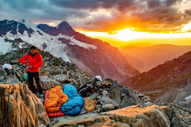 person in red jacket and blue denim jeans sitting on rock near snow covered mountain during